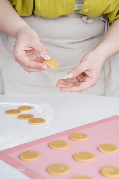Free Crop unrecognizable female confectioner in apron putting raw homemade cookies on baking paper while standing at white table Stock Photo
