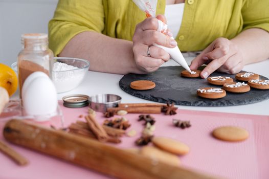 Crop anonymous female confectioner decorating delicious homemade cinnamon cookies with icing sitting at table near flour and cinnamon sticks