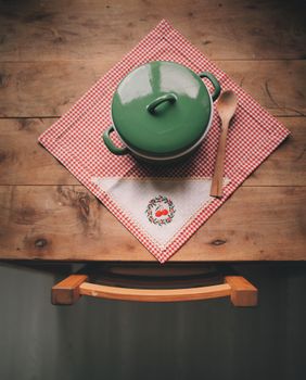 Top view of a rustic wooden table with a green pot, spoon, and red checkered tea towel.