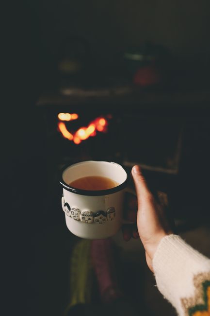 Warmly lit indoor scene featuring a hand holding a cup of tea near a fireplace.