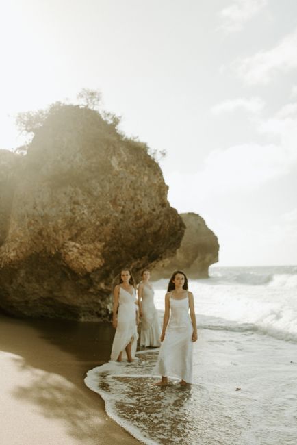 Three women in white dresses walking on a sunny Puerto Rico beach near rocky cliffs.