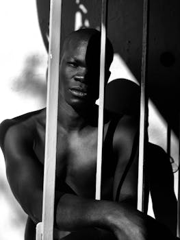 Black and white of confident young African American male with naked torso standing near fence in sunlight and looking at camera