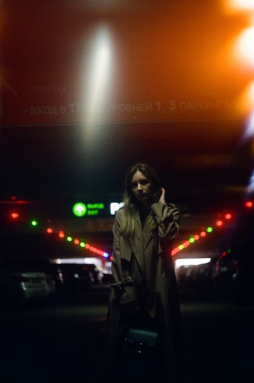 Free Moody portrait of a woman in a trench coat, illuminated by colorful parking lot lights. Stock Photo