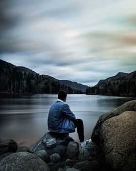 Man in denim jacket sitting by tranquil mountain lake, surrounded by rocks and scenic beauty.