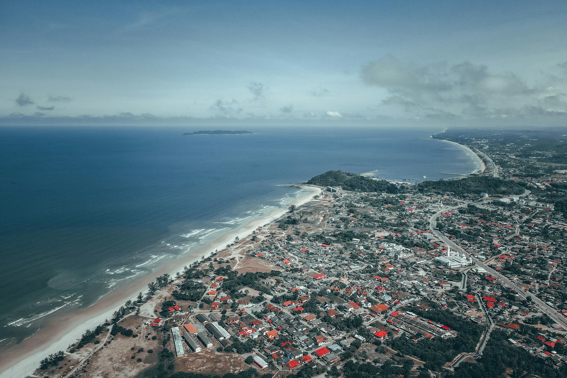 Amazing drone view of residential houses located in city near sandy beach washing by foamy waves of blue ocean against cloudy sky