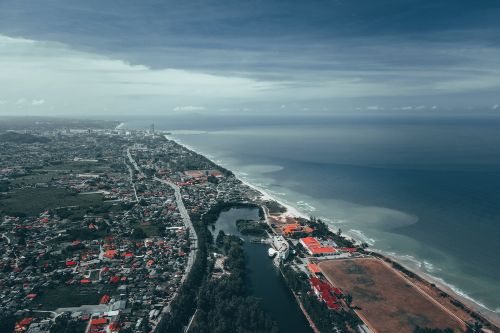 Free Picturesque aerial view of coastal town with residential houses and lush green trees located near sandy beach washing by wavy ocean against cloudy blue sky Stock Photo