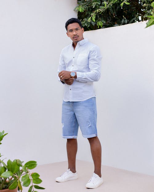 Full body of fashionable young ethnic male with dark hair and eyeglasses in trendy outfit standing in yard of modern house with white walls and looking at camera