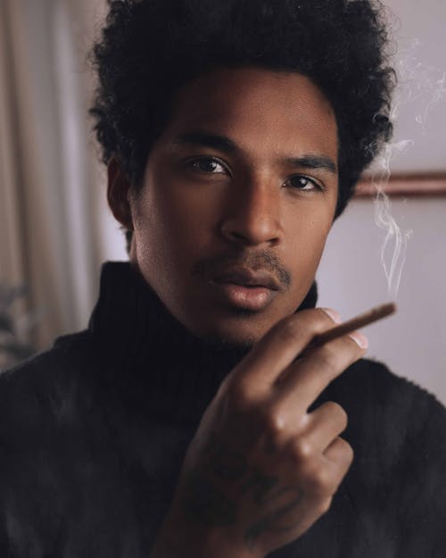 Free Crop pensive African American male with curly dark hair and mustache smoking cigarette and looking at camera in room Stock Photo