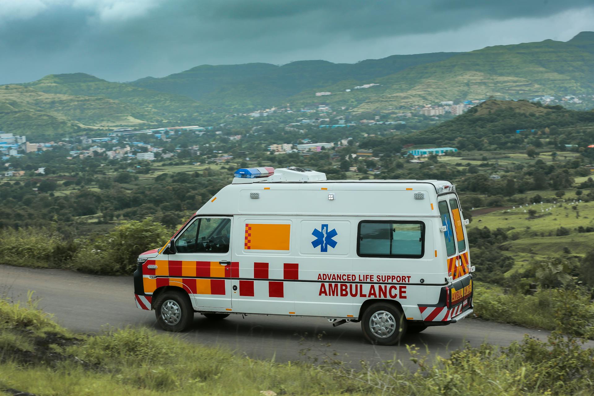 Advanced life support ambulance driving on a scenic road in Pune, India with lush green hills in the background.