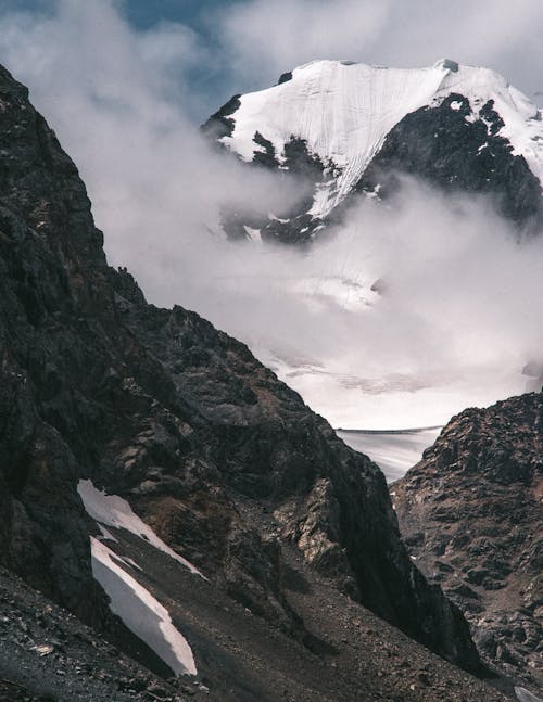 Scenic view of high rough mounts with snow under cloudy sky on sunny day in misty weather