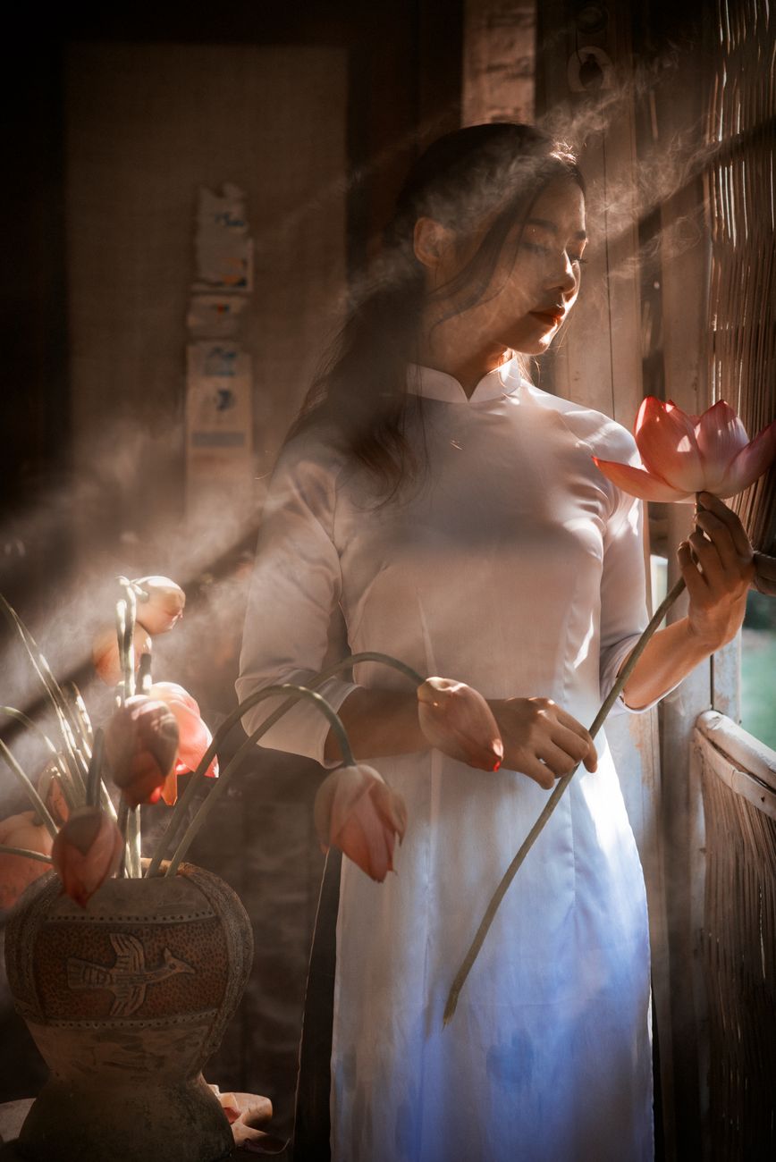 A serene Vietnamese woman in an Ao Dai holding a lotus, captured in soft sunlight indoors.