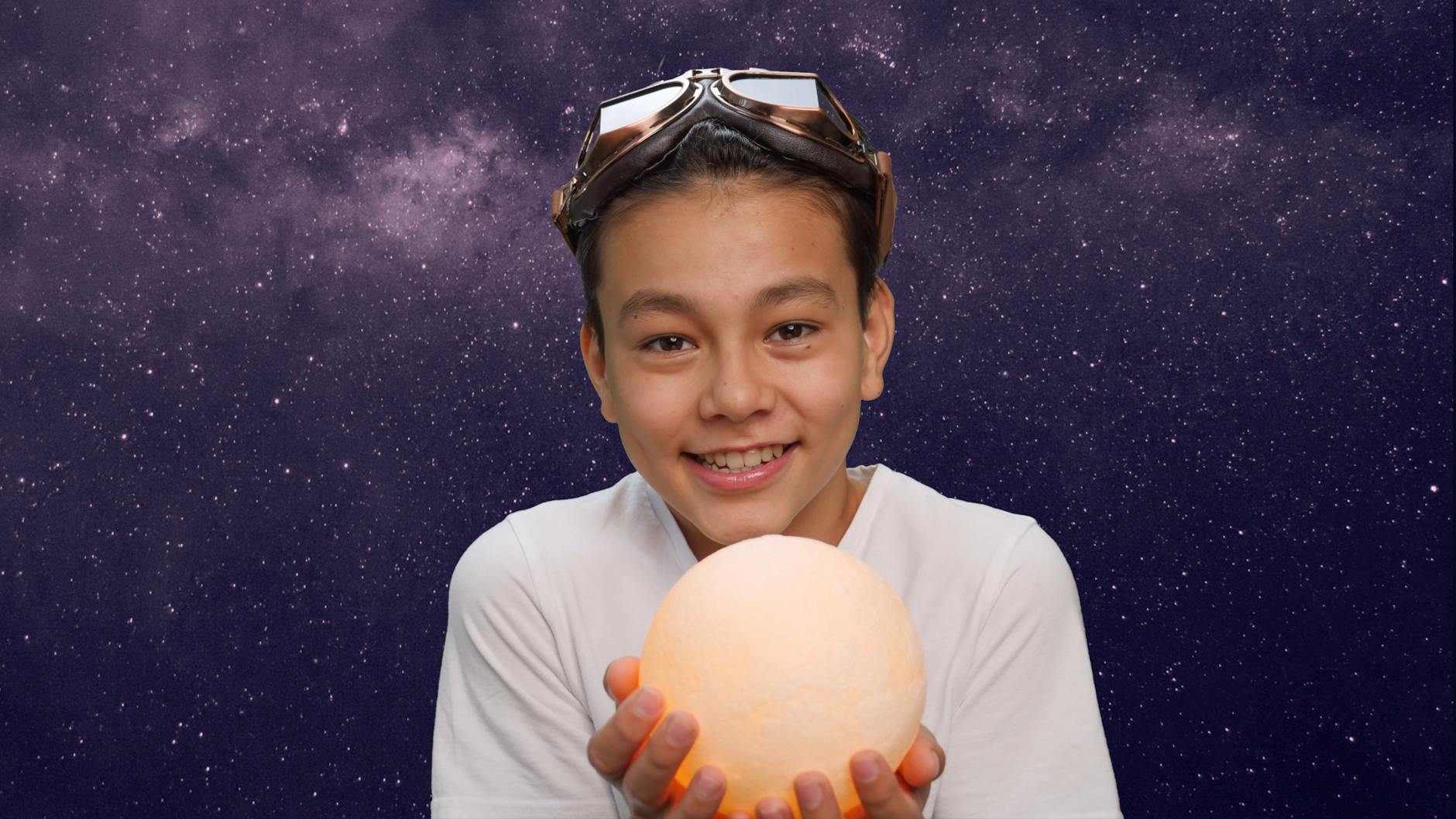 A young boy with goggles holding a glowing sphere against a starry night sky.