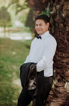 一名身穿正装的亚洲男子在户外倚树微笑，手里拿着夹克