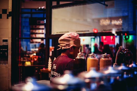 A barista wearing a cap amidst colorful jars in a lively indoor cafe during nighttime.