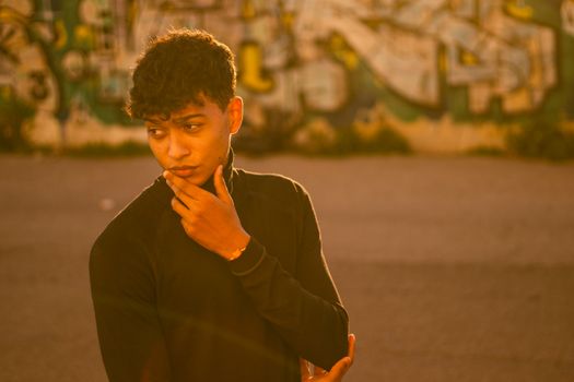 A stylish young man poses against a graffiti backdrop in Valencia, bathed in warm sunset light.