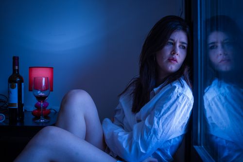 Free A thoughtful woman in white shirt gazing out a window beside a wine bottle. Stock Photo