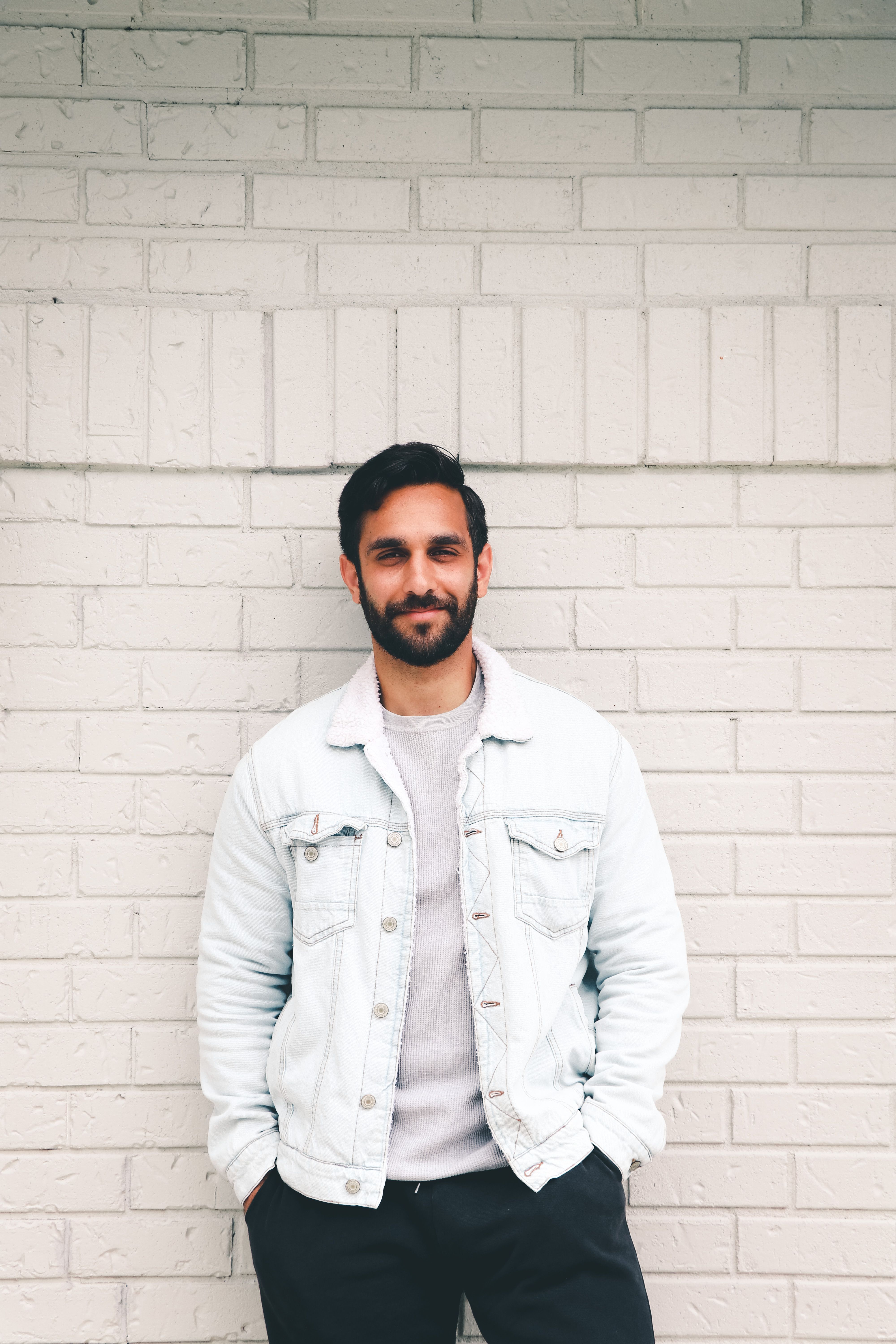 Casual portrait of a bearded man in a denim jacket against a light brick wall.