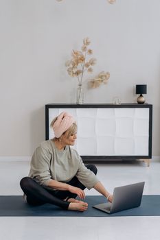 Elderly woman sitting on a yoga mat using a laptop in a stylish living room.
