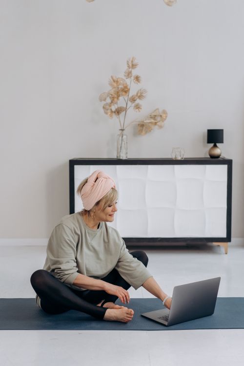 Free Elderly woman sitting on a yoga mat using a laptop in a stylish living room. Stock Photo