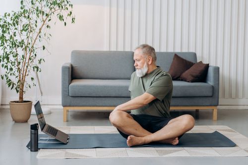 Free Senior man doing yoga on a mat at home, facing a laptop for virtual guidance. Stock Photo
