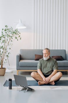 Elderly man engaged in a yoga session at home using a laptop for guidance, sitting on a mat.