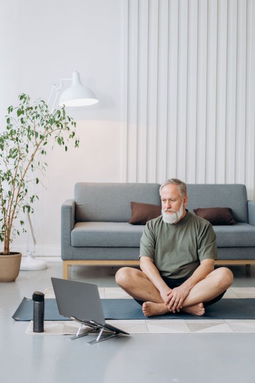 Free Elderly man engaged in a yoga session at home using a laptop for guidance, sitting on a mat. Stock Photo