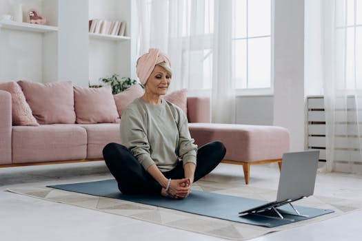 Elderly woman sitting on yoga mat meditating indoors with laptop.