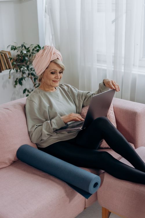 Free Elderly woman using a laptop on a pink sofa with yoga mat nearby, enjoying leisure time at home. Stock Photo