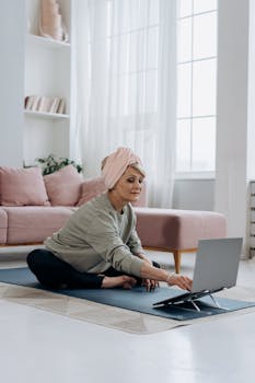 An elderly woman in a living room practicing yoga with a laptop, promoting a healthy lifestyle.