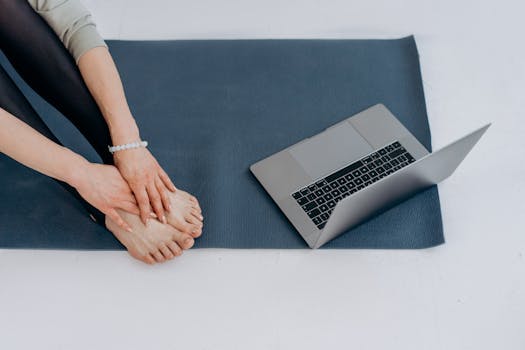Individual on yoga mat practicing stretches alongside a laptop for online fitness classes.