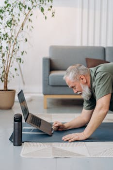 Elderly man engaging in yoga exercise at home using a laptop for guidance.