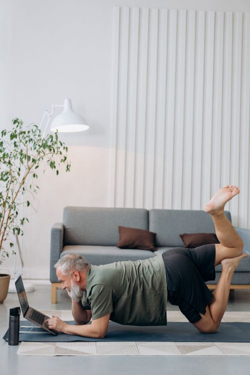 Free Elderly man practicing yoga with a laptop in a modern living room setting. Stock Photo