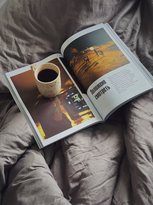 From above cup of aromatic coffee placed on opened magazine on soft cozy bed in morning