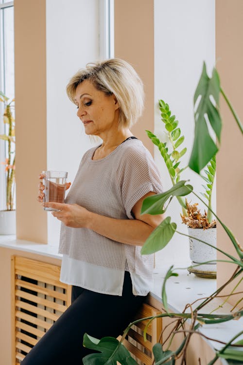 An Elderly Woman in Gray Shirt Leaning on a Windowsill with Green Plants while Holding a Glass of Water