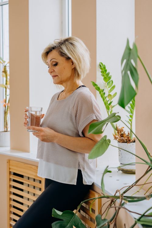 Free A woman in casual attire holds a glass of water while leaning against a windowsill indoors. Stock Photo