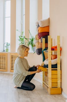 Caucasian woman kneeling indoors organizing yoga props on wooden shelves with natural light.