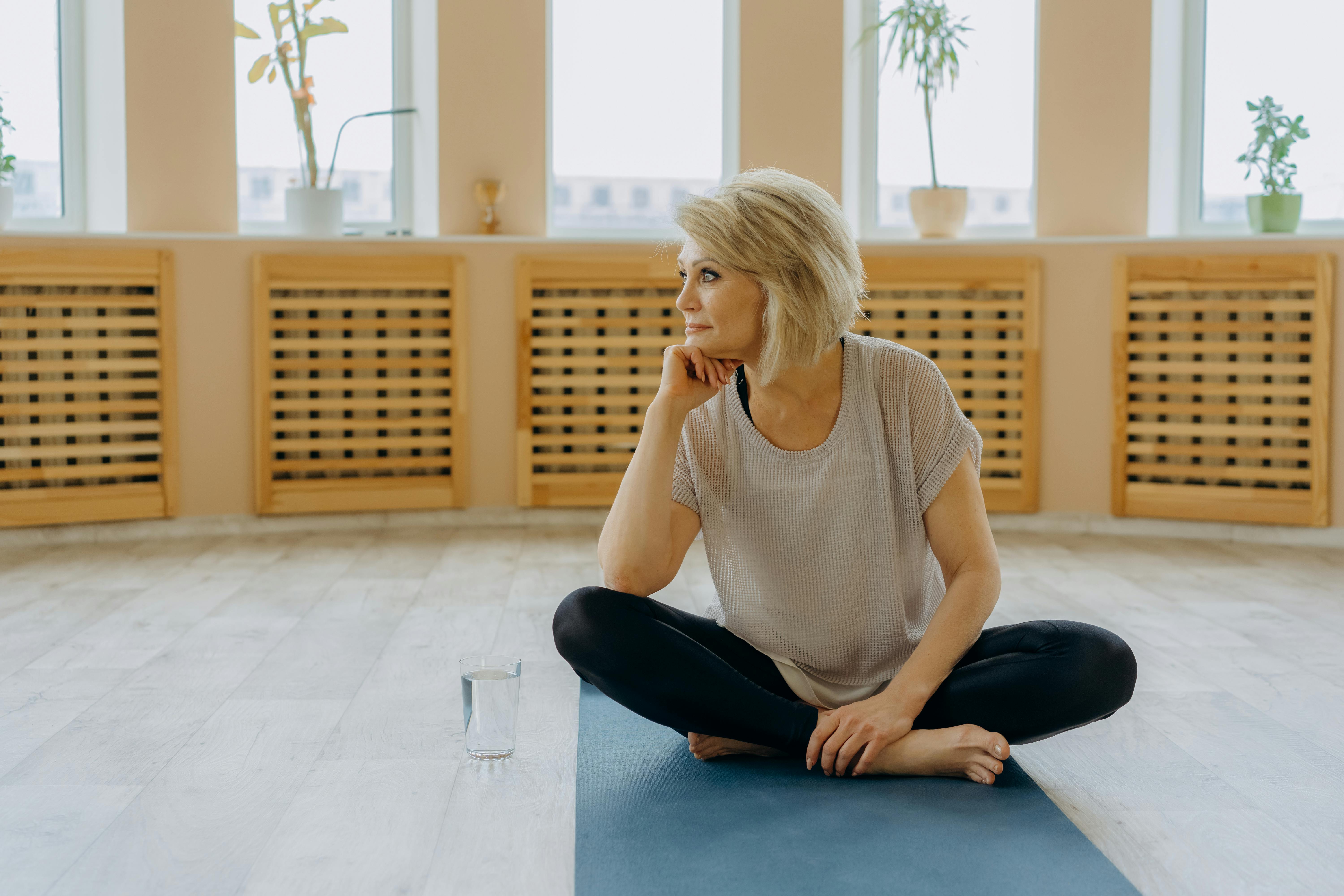 an elderly woman in gray shirt and black leggings sitting on a yoga mat with her hand on her chin