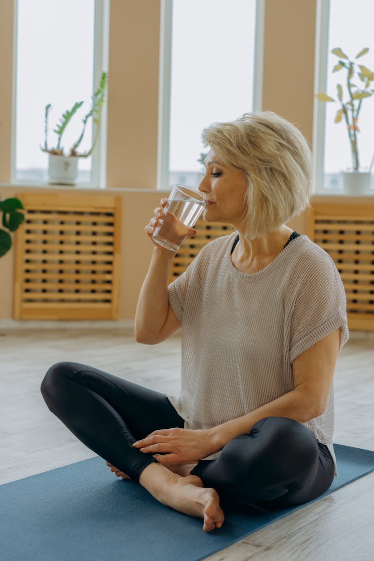 An Elderly Woman In Black Leggings Sitting On Yoga Mat While Drinking Water