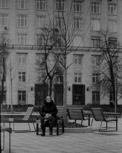 Black and white of aged woman sitting on wooden bench on street against building in city in daylight