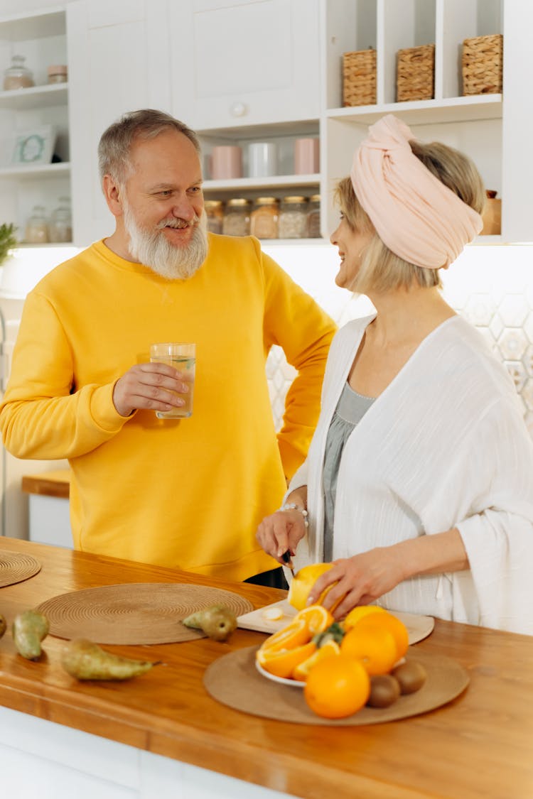 Elderly Man Holding A Glass Of Water Beside Elderly Woman Slicing Orange Fruit