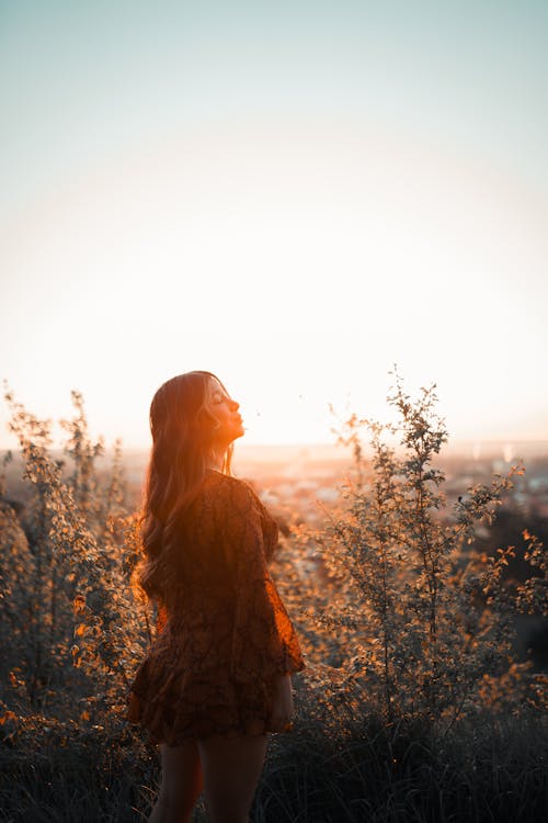 Back view of young female with long wavy hair standing in grassy valley in bright sunset light