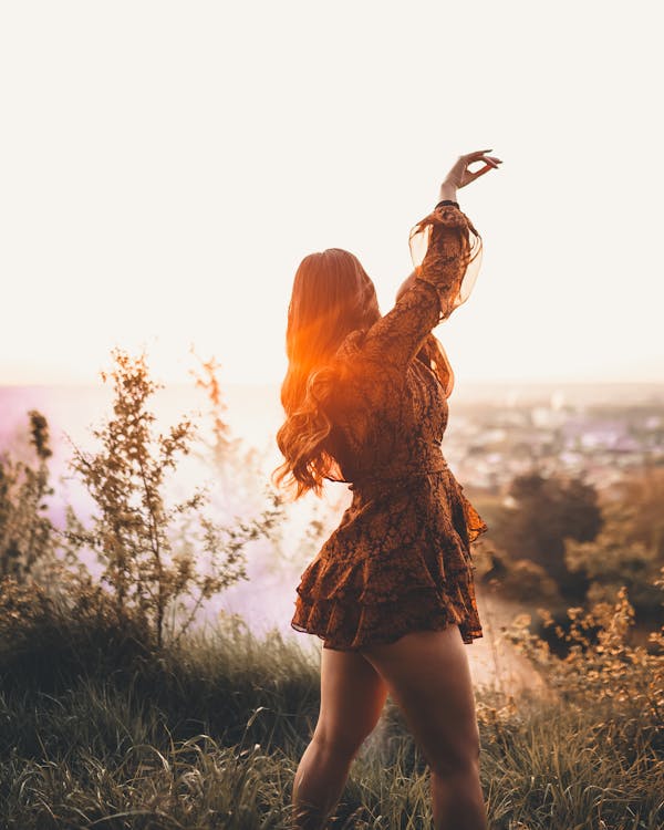 Free Side view of unrecognizable female with long wavy hair raising hand on grassy hill at sundown Stock Photo