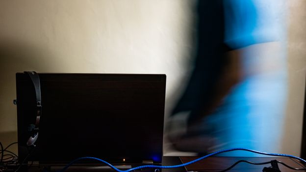 Long exposure of anonymous male in blue t shirt walking toward black computer in light room
