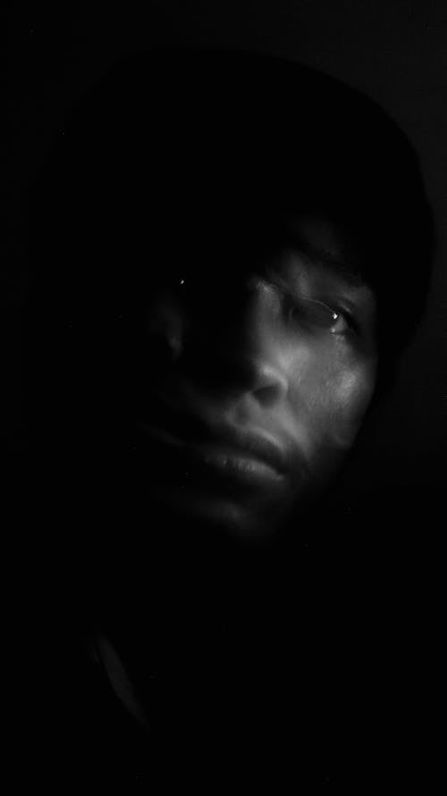 Black and white portrait of thoughtful young Indian male with shadow on face looking away against black background