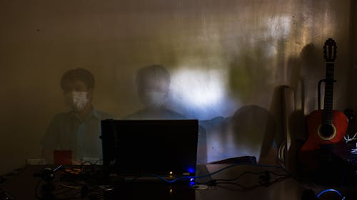 Double exposure of Asian worker in protective mask sitting at table near laptop with wires against guitar during concert