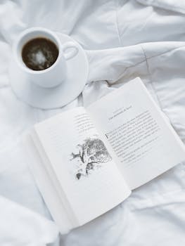 A relaxing scene of a cup of coffee and an open book on white bedding.