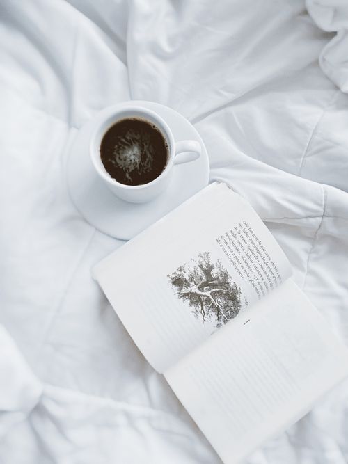 Free A comforting setup of a hot coffee cup and an open book on a white duvet, exuding relaxation. Stock Photo