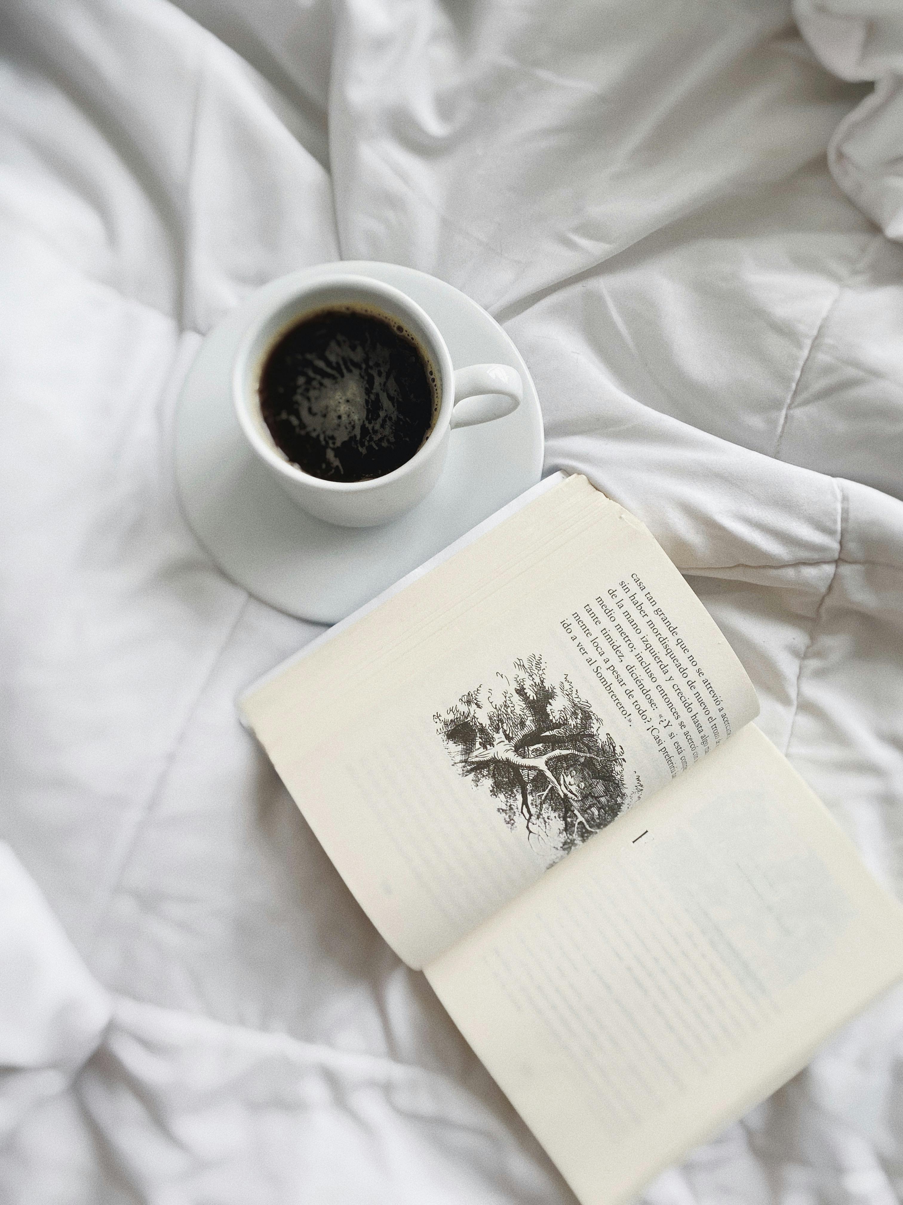 A cup of coffee next to an open book on a white bed, creating a cozy morning atmosphere.