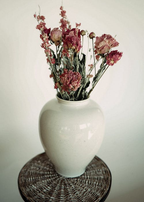 Bunch of dry flowers with thin petals in vase on wicker surface against white wall