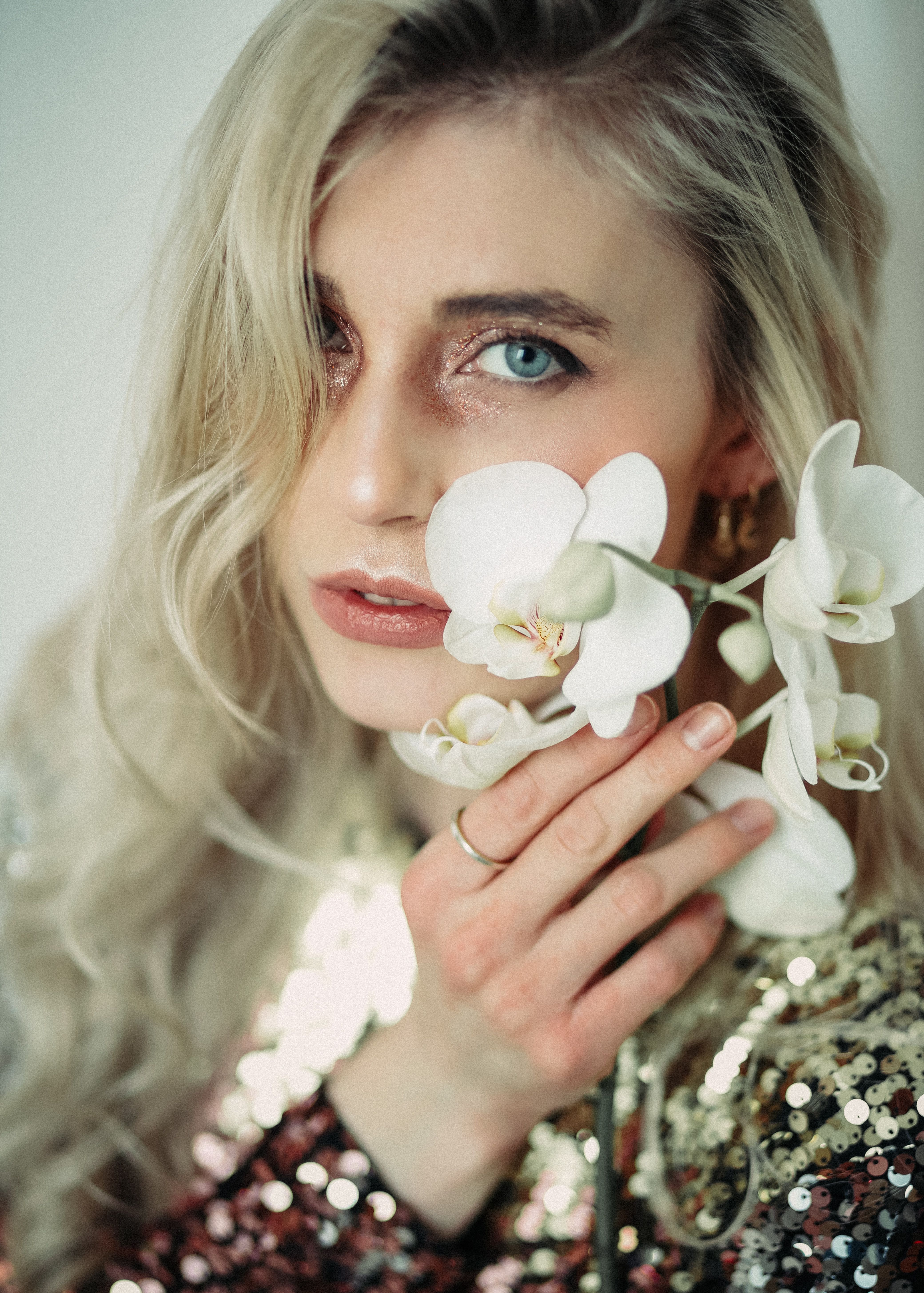 Crop young female with makeup wearing shimmering outfit touching cheek with blossoming flower and looking at camera
